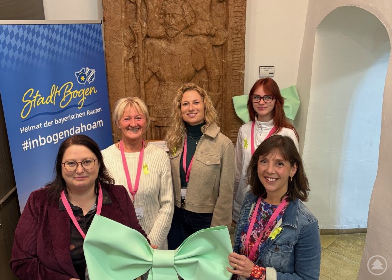 Die Organisatorinnen des Fachtags um Evelyn Jurgasch (vorne links) und Marina Luginger (vorne rechts). Fünf Frauen stehen lächelnd im Eingangsbereich eines Gebäudes. Zwei von ihnen halten eine große grüne Schleife. Im Hintergrund sind ein Banner der Stadt Bogen und eine Wandplastik zu sehen.