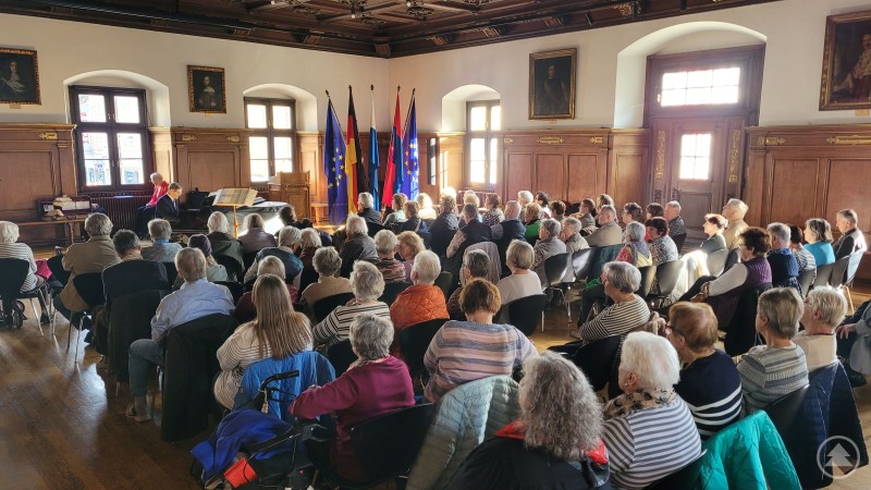 Zahlreiche Besucher genossen das Konzert des Ehepaars Brigitte und Toni Daumerlang im bis auf den letzten Platz gefüllten Historischen Rathaussaal Deggendorf. Eine große Gruppe von Seniorinnen und Senioren sitzt im historischen Rathaussaal Deggendorf und lauscht einem Konzert. Im Vordergrund sind die Zuhörer zu sehen, im Hintergrund die Bühne mit Flügel, Musikern und mehreren aufgestellten Fahnen.