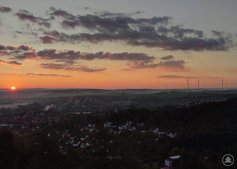 Die Diskussion um regenerative Energien ist stellenweise „nebulös“, was Argumente zu Pro- und Contra betrifft. Die Frage, was Anlagen wie Windräder vor Ort bringen, soll nun mit einer Gemeindebeteiligung klarer geregelt werden. Panoramaaufnahme einer hügeligen Landschaft bei Sonnenaufgang. Im Vordergrund liegen Häuser einer Kleinstadt, im Hintergrund stehen mehrere Windräder unter einem farbig leuchtenden Himmel.