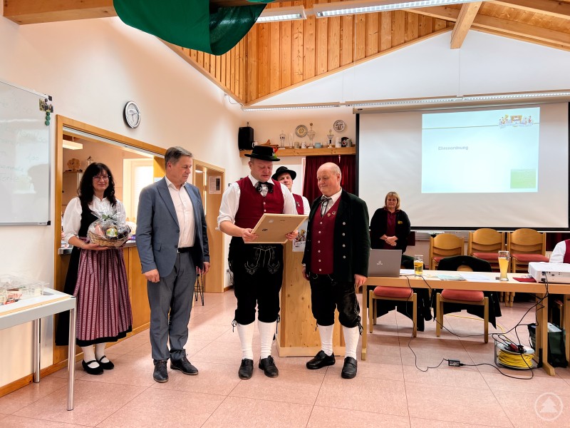 Im Vereinsheim verliest ein Vorstandsmitglied in Tracht eine Urkunde. Daneben steht Franz Behringer ebenfalls in Tracht, weitere Vereinsmitglieder und G&auml;ste verfolgen die Ehrung.