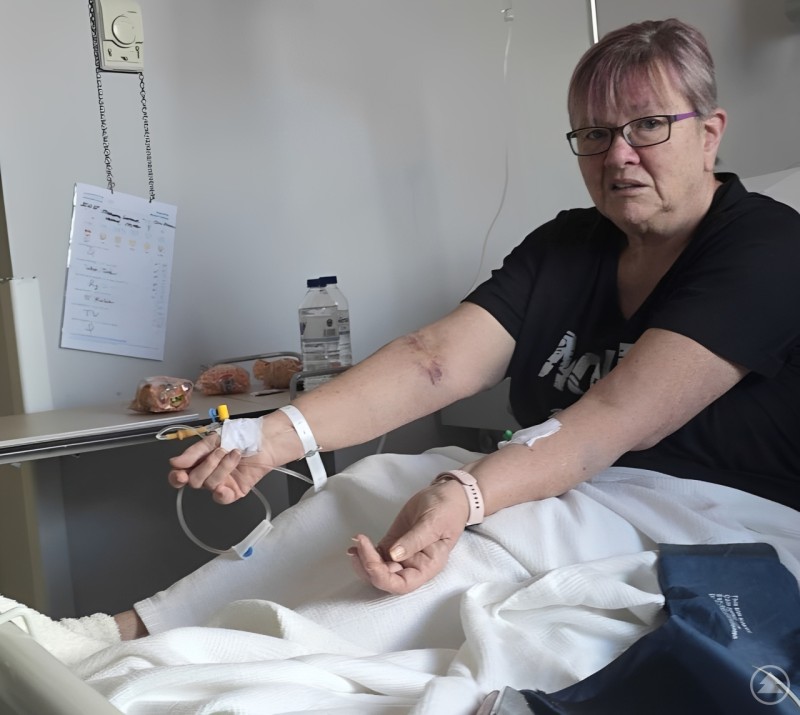 Manuela sitzt im Krankenhausbett und zeigt ihre Arme mit Infusionszug&auml;ngen und Bluterg&uuml;ssen. Sie wirkt ersch&ouml;pft, aber wach und ansprechbar.