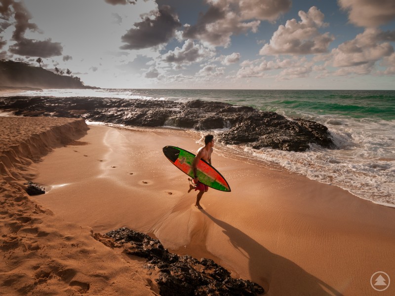 Ein Surfer läuft barfuß mit einem bunten Surfboard über einen sandigen Strand. Im Hintergrund sind dunkle Felsen, Meereswellen und ein wolkiger Himmel zu sehen. Ein Surfer mit Board am Strand von Hawaii vor felsiger Küste und türkisfarbenem Meer.