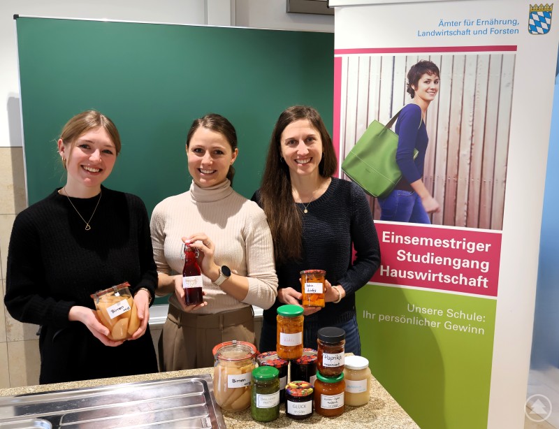 Die Studierenden Ann-Cathrin Gottinger und Claudia Murr haben im Unterrichtsfach „Küchenpraxis“ verschiedene Formen der Haltbarmachung von Lebensmitteln kennen gelernt. Lehrerin Vanessa Handl (li) präsentiert mit ihnen einen Teil der Ergebnisse. Drei junge Frauen stehen hinter einer Arbeitsfläche mit zahlreichen Einmachgläsern und selbstgemachten Vorräten. Im Hintergrund ein Aufsteller zum einsemestrigen Studiengang Hauswirtschaft und eine grüne Tafel.