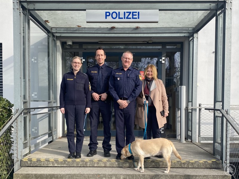 Das Foto zeigt die eingeladene Besucherin zusammen mit (von links) Polizeihauptkommissarin Johanna Vogl, „Helfer in Not“ Polizeioberkommissar Michael Fleischmann, Dienststellenleiter Polizeidirektor Markus Völkl Vor dem Eingang der Polizei stehen drei Polizeibeamte und eine Besucherin, vorne läuft ein Hund über die Treppe. Über ihnen ist das Schild POLIZEI zu sehen.