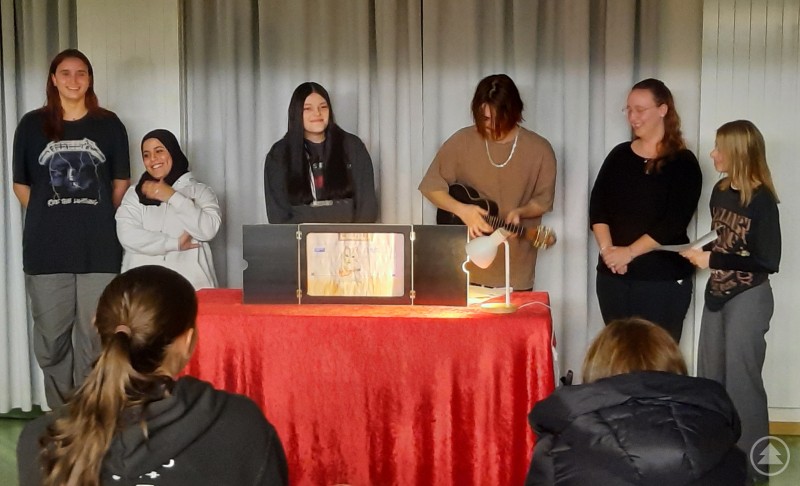 Mehrere Sch&uuml;lerinnen und Sch&uuml;ler stehen auf einer kleinen B&uuml;hne und pr&auml;sentieren eine kreative Darbietung mit Musik und Erz&auml;hltheater vor einem Publikum.