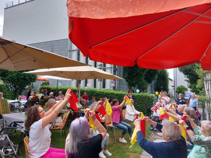 Die Bewohnerinnen und Bewohner des Seniorenstifts zeigten beim Sitztanz große Begeisterung und wurden dabei von vielen Gästen unterstützt. Bewohnerinnen und Besucher des Seniorenstifts Heiliggeist nehmen an einer fröhlichen Gymnastikrunde im Garten teil. Sie sitzen unter großen Sonnenschirmen und schwenken bunte Tücher.