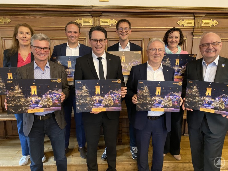 Freuen sich über den neuen Rotary-Adventskalender 2025 (vorn v.l.): Landrat Bernd Sibler, Oberbürgermeister Dr. Christian Moser, Rotary-Präsident Hermann Peter und Distrikt Governor Ulli Kersting; hinten v.l.: Brigitte Wallstabe-Watermann, Maximilian Roth, Markus Lantermann und Christiane Lantermann. Mehrere Personen stehen in einem holzvertäfelten Raum und halten den neuen Rotary-Adventskalender 2025 in den Händen, lächelnd in die Kamera.