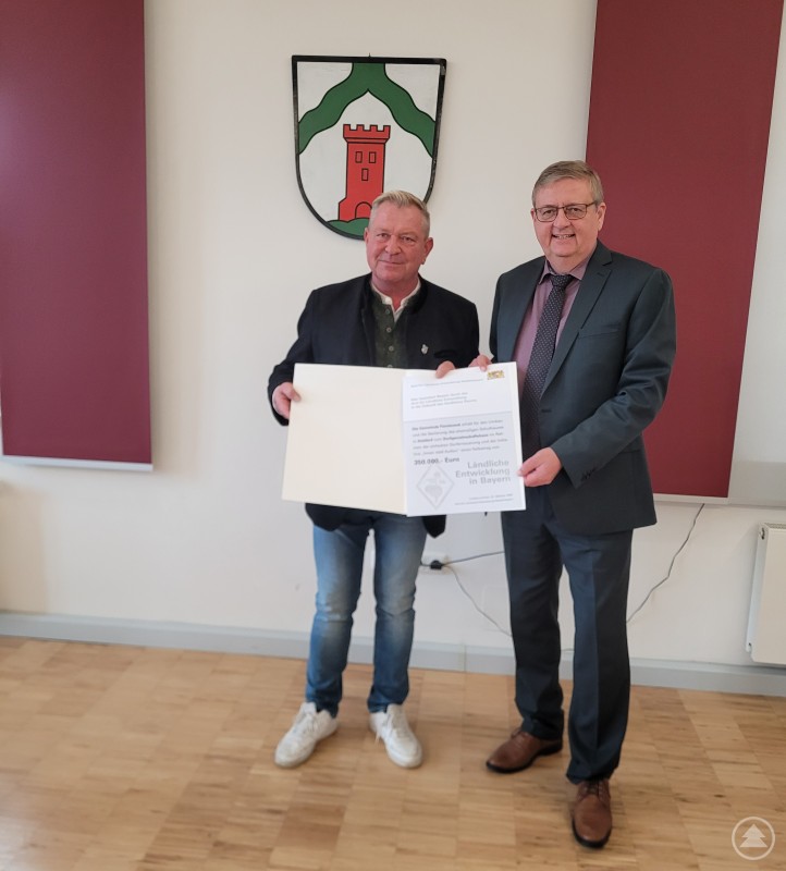 Übergabe der Auszahlungsurkunde: Bürgermeister Alexander Pieringer (links) und Hans-Peter Schmucker, Leiter des ALE Niederbayern. Zwei Männer stehen vor dem Wappen der Gemeinde Fürsteneck. Sie halten gemeinsam eine Förderurkunde des Amts für Ländliche Entwicklung Niederbayern in den Händen und lächeln in die Kamera.