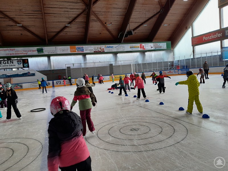 Schüler trainieren verschieden Fahrtechniken an acht unterschiedlichen Stationen. Kinder mit bunten Helmen und Winterkleidung üben auf einer Eisfläche verschiedene Fahrtechniken. In der Eishalle sind mehrere Stationen mit Markierungshütchen aufgebaut, während einige Erwachsene das Geschehen beaufsichtigen.