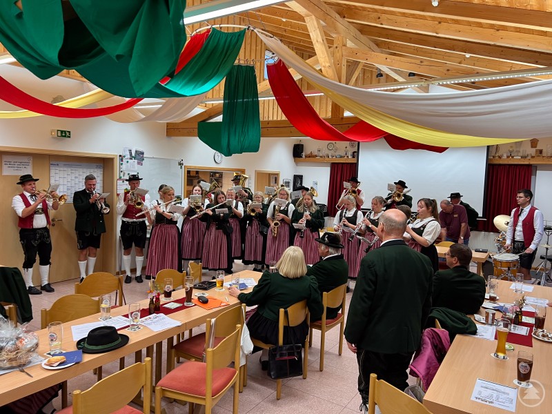 Die Hofmarkkapelle Haus i. Wald spielt in Tracht im Vereinsheim. Zahlreiche Musikerinnen und Musiker stehen mit Instrumenten vor den sitzenden G&auml;sten.