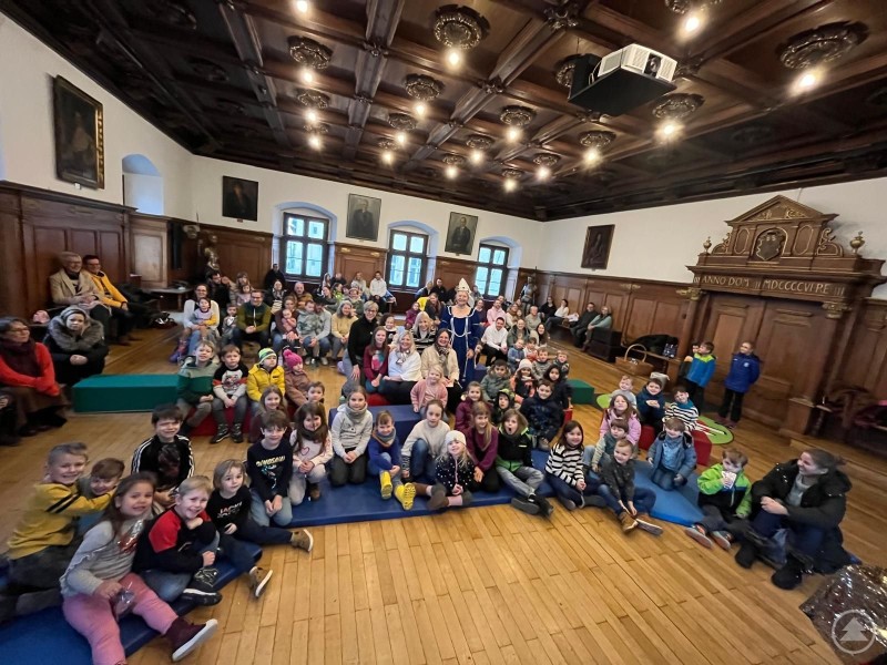 Die Gäste des zauberhaften Nachmittags mit den Organisatoren in der Mitte des Bildes. Viele Kinder und Erwachsene sitzen und stehen in einem historischen Saal mit Holzvertäfelung und verziertem Kassettendach. In der Mitte des Raums sitzen zahlreiche Kinder auf Matten und Bänken, umgeben von Eltern und Veranstaltern, im Hintergrund das kunstvoll verzierte Portal des Alten Rathauses.