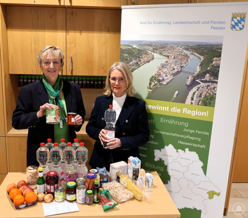Abteilungsleiterin Sieglinde Preuß (li) und Monika Rothbächer, Lehrerin im Unterrichtsfach „Ernährung und Lebensmittel“ (re), zeigen eine Auswahl an Lebensmitteln, die den Bedarf eines Zweipersonenhaushaltes für drei Tage abdecken kann. Bei größeren Familien muss dieser entsprechend hochgerechnet werden. Zwei Frauen stehen hinter einem Tisch mit zahlreichen haltbaren Lebensmitteln, darunter Wasserflaschen, Konserven, Nudeln und Obst. Im Hintergrund ein Aufsteller des Amts für Ernährung, Landwirtschaft und Forsten Passau.