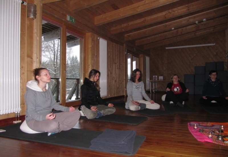 Mehrere Frauen sitzen auf Yogamatten in einem Holzraum im Schneelandschaft-Ambiente und meditieren mit geschlossenen Augen. Vorne liegen Decken, rechts sind Kerzen und Tücher als ruhige Dekoration zu sehen.
