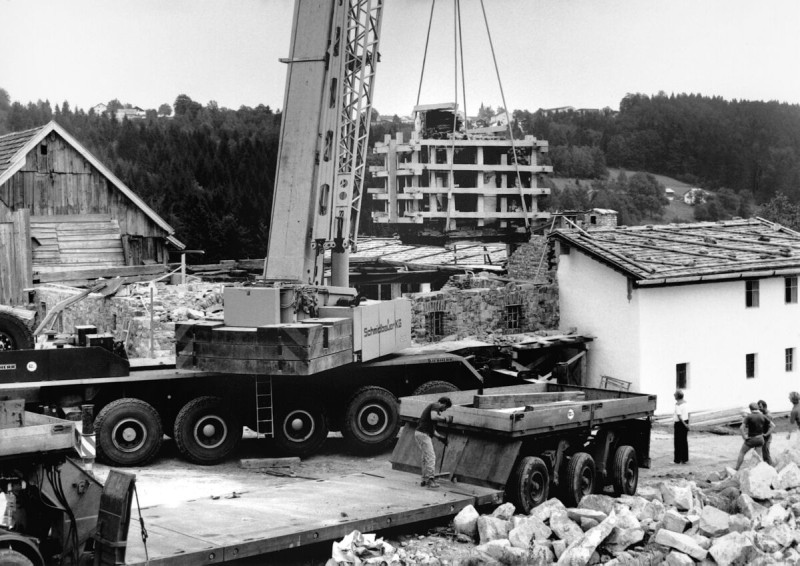 Abbau der Rauchkuchl und Übertragung in einem Stück Schwarzweißaufnahme eines großen Baukrans, der ein schweres Betonteil hebt. Im Hintergrund sind Baustellenstrukturen und bewaldete Hügel zu sehen – vermutlich eine Szene aus dem industriellen oder infrastrukturellen Aufbau der Nachkriegszeit.