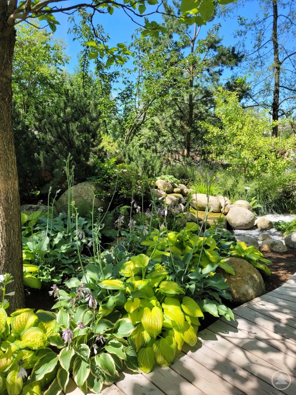 Die Themengärten bieten Rückzugsorte und Raum zur Entfaltung im Grünen. Ein idyllischer Garten mit üppigem Grün, schattenspendenden Bäumen und einem Bachlauf, eingerahmt von Zierpflanzen und großen Steinen.