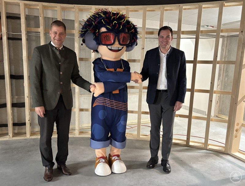 Drei Monate vor der Eröffnung: Bürgermeister Dr. Olaf Heinrich (links) und Landrat Sebastian Gruber (rechts) mit Maskottchen TonY im TonYversum-Gebäude, wo ab 25. April die Bayerische Landesausstellung 2026 „Musik in Bayern“ stattfindet. Zwei Männer in Anzug bzw. Tracht stehen gemeinsam mit einem großen Maskottchen mit blauen Haaren und roter Brille in einem modernen, im Rohbau befindlichen Ausstellungsraum. Das Maskottchen schüttelt dem rechten Mann die Hand.