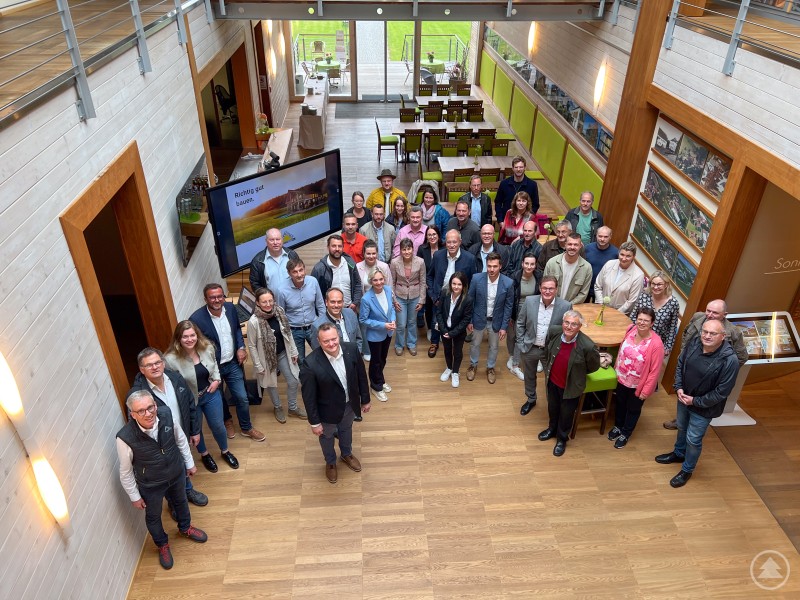 Mitglieder des Ilzer Land Unternehmernetzwerks, der Wirtschaftsjunioren Passau und der Mittelstandsunion Rottal-Inn zusammen mit den Geschäftsführern (Hr. Gotthard Sonnleitner Foto Mitte) und Mitarbeitern von Sonnleitner Eine große Gruppe von Unternehmerinnen und Unternehmern steht in einem modernen, lichtdurchfluteten Ausstellungsraum in Holzoptik, aufgenommen von oben. Im Hintergrund ist ein Bildschirm mit der Aufschrift „Richtig gut bauen.“ zu sehen.