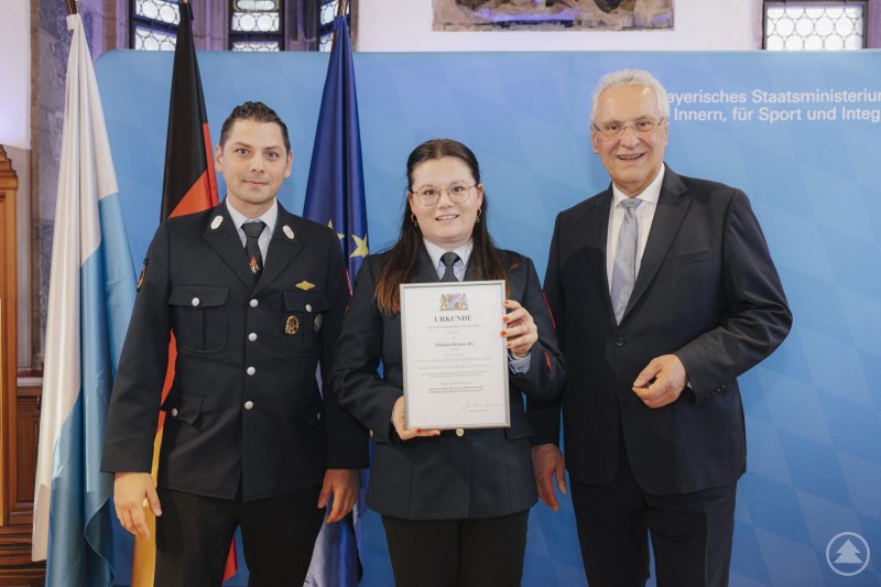 Uli Hurzlmeier und Steffi Graf (beide Thomas-Krenn.AG) mit Staatsminister Joachim Herrmann Zwei Feuerwehrangehörige der Thomas-Krenn.AG nehmen die Auszeichnung „Ehrenamtsfreundlicher Betrieb“ von Staatsminister Joachim Herrmann entgegen. Im Hintergrund sind die Flaggen von Bayern, Deutschland und der EU zu sehen.