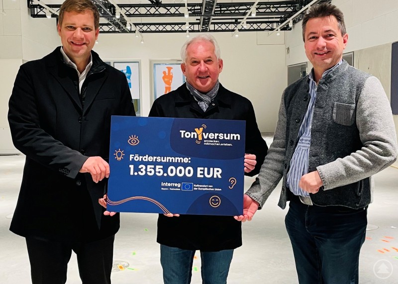 Bürgermeister Dr. Olaf Heinrich (Mitte) freut sich gemeinsam mit Projektbeteiligten über die INTERREG-Förderzusage für das Science Center TonYversum in Freyung. Drei Männer stehen in einem hellen Ausstellungsraum und halten gemeinsam ein großes Förderscheck-Plakat mit der Aufschrift TonYversum Freyung und dem Betrag 1.355.000 Euro.