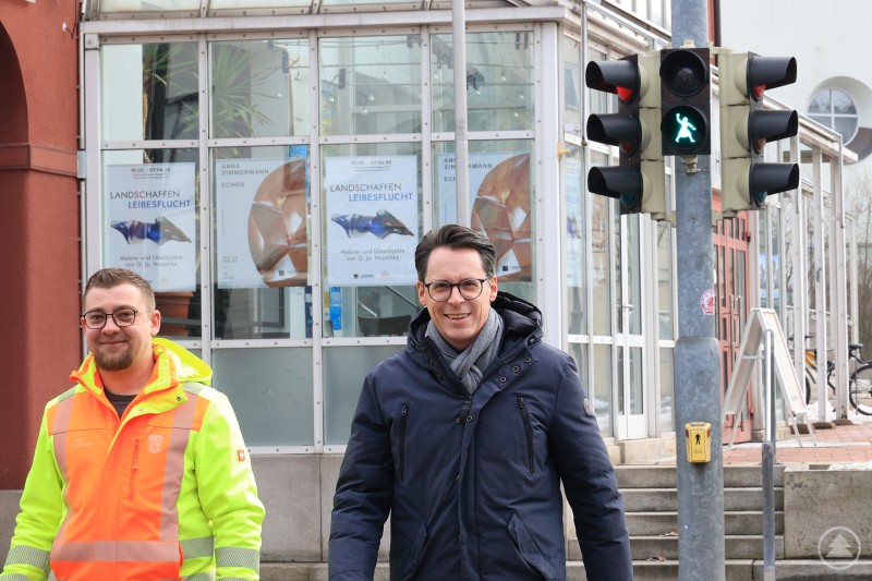 Florian Geiger (Stadt Deggendorf, links) und Oberbürgermeister Dr. Moser testen die neue Fußgängerampel mit Knödelwerferin-Motiv im Östlichen Stadtgraben. Zwei Männer stehen lächelnd vor einer Fußgängerampel mit Knödelwerferin-Motiv, einer trägt eine Warnjacke. Im Hintergrund sind Glasfenster und Plakate einer Ausstellung zu sehen.