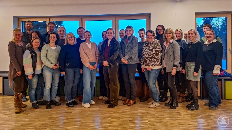 Beim Jahrestreffen der Nationalpark-Schulen im Jugendwaldheim gab es viele Gelegenheiten zum gegenseitigen Austausch. Eine große Gruppe von Erwachsenen steht in einem hellen Raum mit Holzfußboden und großen Fenstern. Die Personen lächeln in die Kamera und wirken entspannt, einige stehen eng beisammen, als Gruppenfoto.