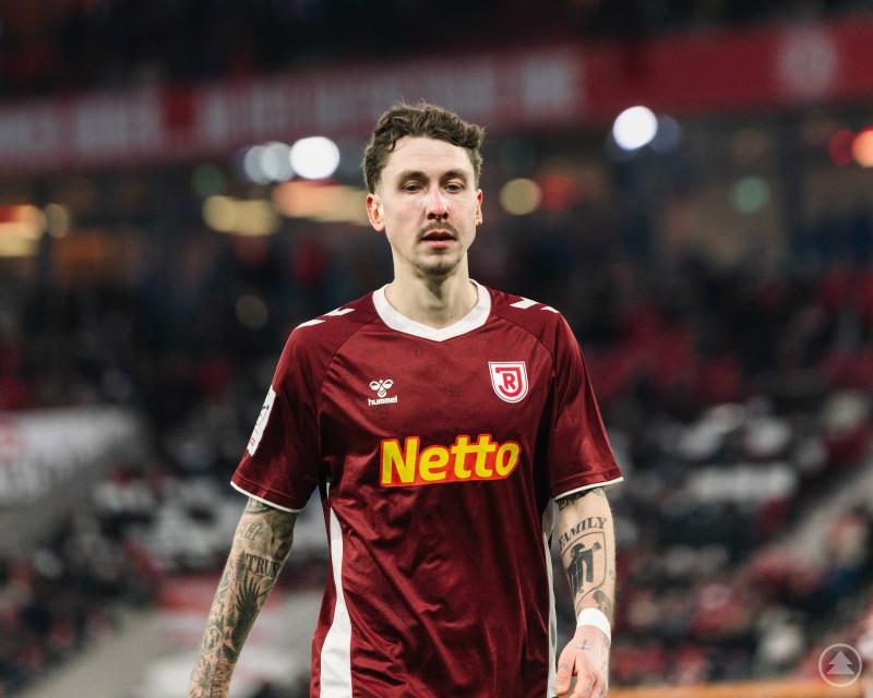 Adrian Fein analysierte nach dem Spiel die knappe Niederlage bei Rot Weiss Essen. Ein Spieler des SSV Jahn Regensburg im bordeauxroten Trikot blickt nachdenklich über den Platz, im Hintergrund verschwommene Stadiontribünen.