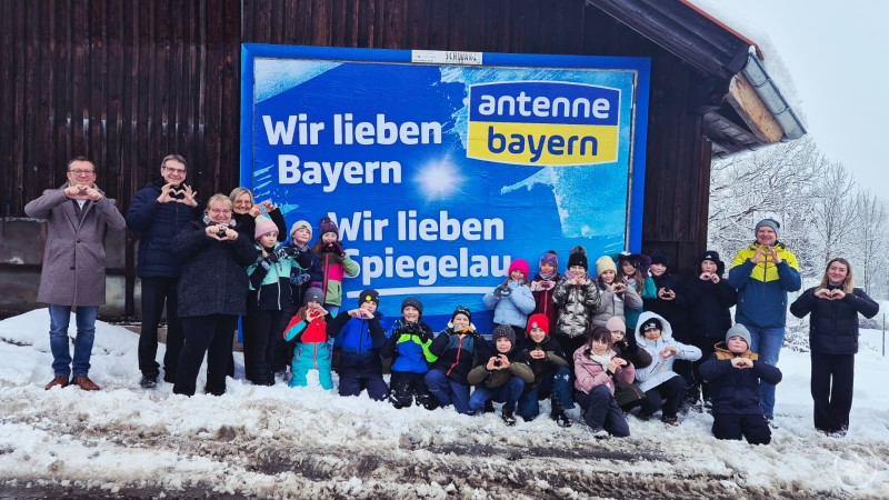 v. l. Günther Nama (2. Bürgermeister), Karlheinz Roth (1. Bürgermeister), Corinna Stibale (Elternbeiratsvorsitzende), Petra Strohmaier (stellvertretende Schulleiterin), Klasse 4a, Michael Prager (Klassenlehrer) und Marie Schreiner. Eine große Gruppe aus Kindern und Erwachsenen steht im Schnee vor einer Holzwand mit einem großen Plakat von Antenne Bayern. Alle machen mit den Händen ein Herzzeichen. Im Hintergrund Bäume und Winterlandschaft.