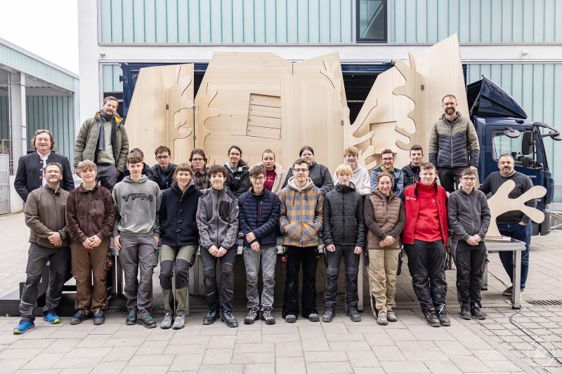 Schülerinnen und Schüler der Landshuter Berufsschule 1 (BS1), Fachbereich Holztechnik, vor der Bühne des fahrenden Theaters; Schulleiter BS1 Reinhold Ostermaier (li., 2. Reihe), Ole Wegener (li., 1. Reihe), BS1, Dr. Laurenz Schulz, Projektleiter KULURmobil (li. oben), Josef Hundhammer (re. oben) und Franz Pledl (re.), BS1 Eine große Gruppe von Schülerinnen und Schülern sowie Lehrkräften und Verantwortlichen steht im Hof vor einem Lkw mit aufgebauter, holzfarbenen Bühnenkulisse. Die Kulissenteile sind großformatig und zeigen stilisierte Formen, passend für das fahrende Theater KULTURmobil.