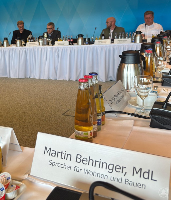 Klausureinblick in Gmund: Martin Behringer, MdL, im Austausch mit Kabinettsmitgliedern über die Zukunft der Wohnbauförderung. Ein Konferenztisch mit mehreren Getränkeflaschen, Mikrofonen und Namensschildern, darunter deutlich sichtbar „Martin Behringer, MdL“. Im Hintergrund sitzen weitere Teilnehmer einer politischen Klausur, darunter auch Vertreter mit dem Titel „Staatsminister“.