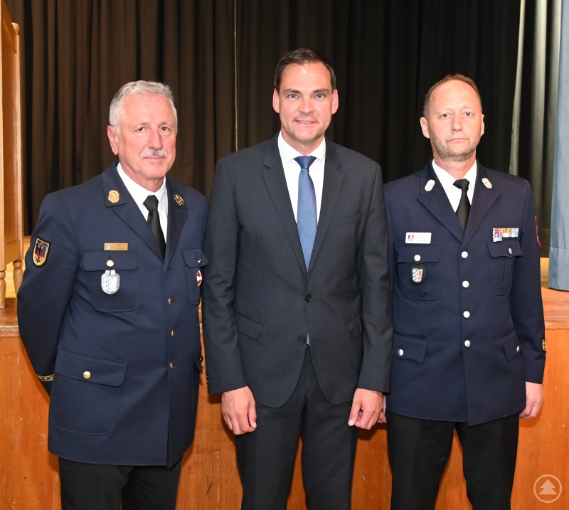 Ab November wird Martin Bauer (rechts) das Amt des Kreisbrandrates von Norbert Süß (links) übernehmen. Norbert Süß, Sebastian Gruber und Martin Bauer stehen nebeneinander vor einem Vorhang – zwei in Feuerwehruniform, einer im Anzug.