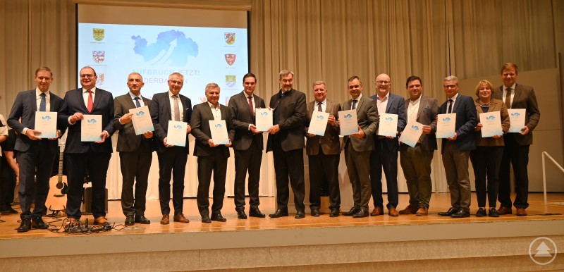 Ministerpräsident Dr. Markus Söder (Mitte) nahm den Maßnahmenkatalog „Aufbruch jetzt! Niederbayern“ in Vilshofen entgegen. Gemeinsam mit den Landräten und Oberbürgermeistern präsentierte er den Zukunftsplan für eine starke Region. Eine Gruppe von Landräten und Oberbürgermeistern steht gemeinsam auf einer Bühne in einem Festsaal. Sie halten den Maßnahmenkatalog des Projekts „Aufbruch jetzt! Niederbayern“ in den Händen und lächeln in die Kamera. Im Hintergrund ist eine Leinwand mit dem Projektlogo zu sehen.