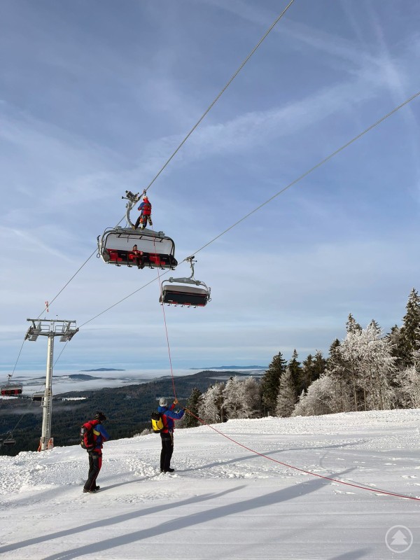Rettung aus der Höhe: Mit Seiltechnik werden Personen sicher aus der Sesselbahn geborgen. Bergwacht-Einsatzkräfte seilen sich bei einer Übung zu einer Sessellift-Gondel ab, während weitere Teammitglieder am Boden sichern. Verschneite Berge und Wälder erstrecken sich im Hintergrund.