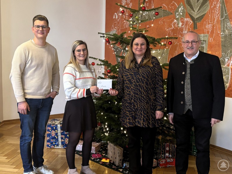 Die Klassensprecher Christoph Lechner und Selina Wagner überreichen Ingrid Gooss, Vertreterin des Kinderhospizes Haus ANNA, eine Spende in Höhe von 1.620 Euro in Beisein von Helmut Ramesberger, Behördenleiter AELF Passau. Vier Personen stehen vor einem geschmückten Weihnachtsbaum in einem festlichen Raum. Zwei junge Erwachsene halten einen Umschlag mit der Spendensumme in den Händen. Daneben stehen eine Vertreterin des Kinderhospizes sowie ein Vertreter der Schulleitung. Im Hintergrund sind Geschenke unter dem Weihnachtsbaum zu sehen.