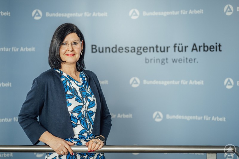 Eva Maria Kelch, Vorsitzende der Geschäftsführung der Agentur für Arbeit Passau Porträt einer Frau mit dunklem Haar und Brille vor einem Hintergrund mit dem Logo der Bundesagentur für Arbeit. Sie steht an einem Geländer und blickt in die Kamera.