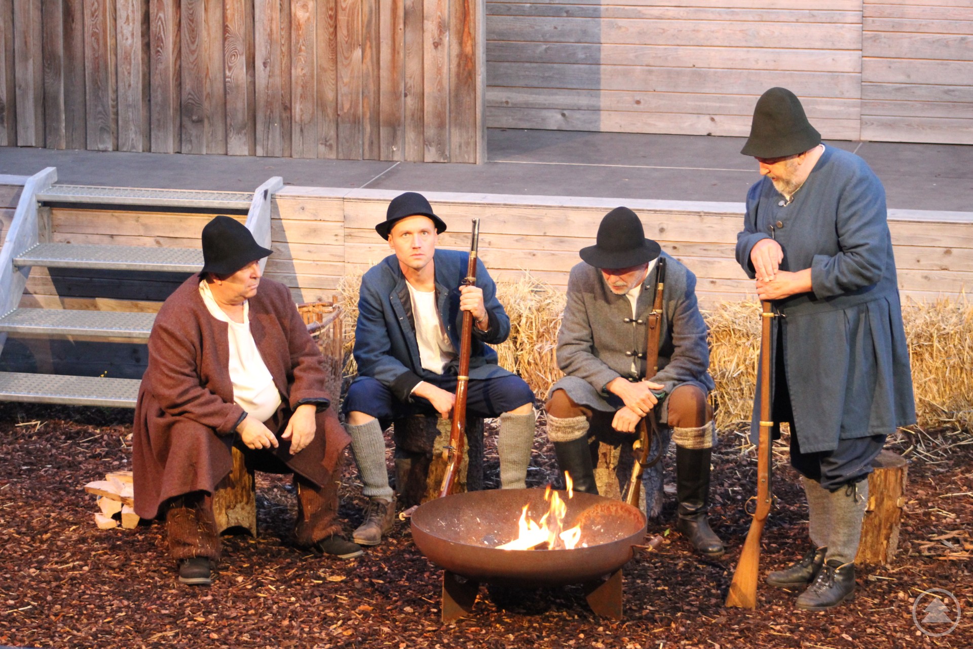 Die Szenen der beratschlagenden Bauern am Lagerfeuer – im Zwiespalt von Für und Wider – sind der rote Faden im Freilichtspiel Auf dem Bild sind vier Männer in historischer Kleidung zu sehen, die um ein brennendes Lagerfeuer sitzen bzw. stehen. Sie tragen einfache Bauerntrachten mit Hüten und halten Musketen in den Händen. Die Szene spielt offensichtlich im Rahmen eines historischen Freilichtspiels – vermutlich aus dem 18. Jahrhundert – vor einer Kulisse aus Holz. Die dargestellte Stimmung ist nachdenklich und ernst, was gut zur Bildunterschrift passt: