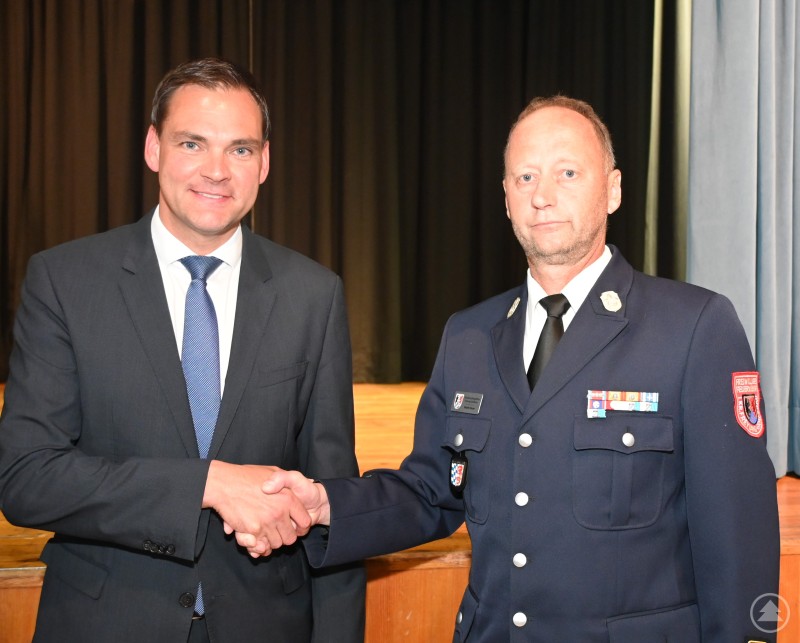 Landrat Sebastian Gruber gratuliert Martin Bauer per Handschlag zu seiner Wahl, beide stehen im Anzug bzw. in Feuerwehruniform vor dunklem Bühnenvorhang.