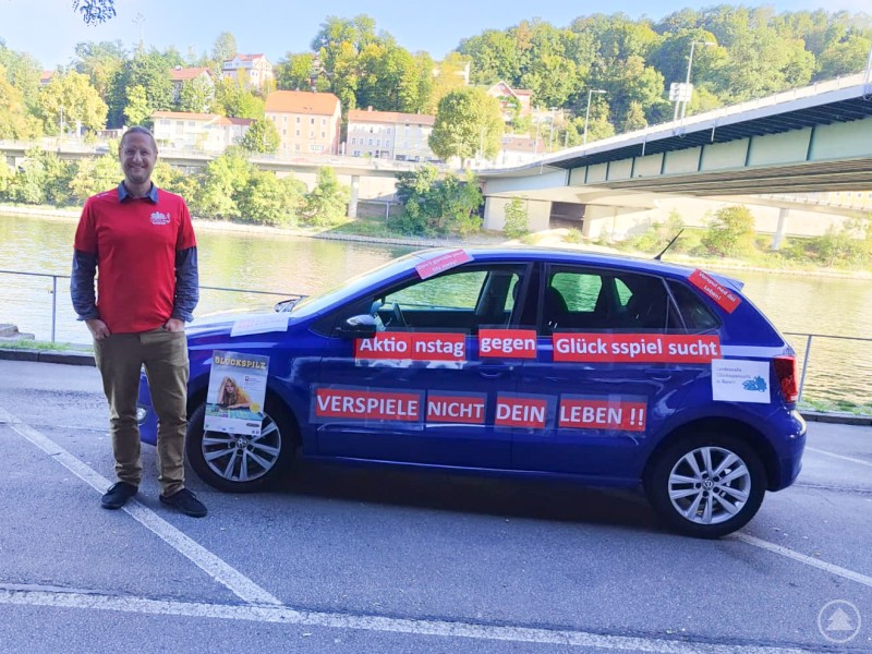 Suchtberater Klaas Spettmann von der Caritas Passau wird am Aktionstag im Landkreis unterwegs sein, um auf die Gefahren von Glücksspielsucht hinzuweisen. Ein Mann in rotem Caritas-Shirt steht neben einem blau beklebten Auto, das mit auffälligen Slogans gegen Glücksspielsucht beschriftet ist. Im Hintergrund ist eine Brücke über einen Fluss zu sehen.