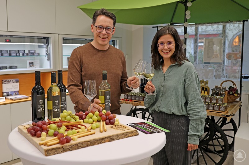 Oberbürgermeister Christian Moser und Organisatorin Selina Haimerl freuen sich auf die bevorstehende Premiere des neuen Deggendorfer Genussevents „Weinloben“. Ein Mann und eine Frau stoßen mit einem Glas Weißwein an. Auf dem Tisch vor ihnen stehen Weinflaschen mit der Aufschrift „Stadtwein Deggendorf“, dazu Trauben, Käsewürfel und Brotstangen. Im Hintergrund ist ein Verkaufsstand mit regionalen Produkten zu sehen.