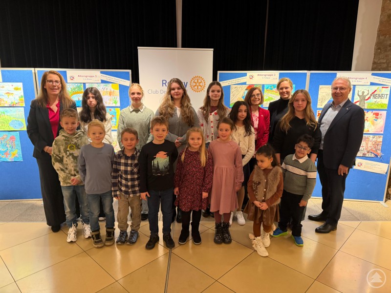 Die Preisträgerinnen und Preisträger des Rotary-Malwettbewerbs 2025 mit Jury und Präsident Hermann Peter (v.r.) Gruppe von Kindern, Jugendlichen und Erwachsenen steht vor blauen Stellwänden mit Wettbewerbsbildern. Alle lächeln, einige sind feierlich gekleidet, im Hintergrund Rotary-Banner.