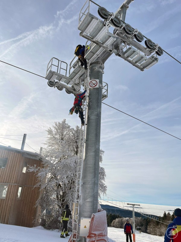 Mehrere Einsatzkräfte der Bergwacht und Feuerwehr klettern bei einer Bergeübung an einem vereisten Sesselbahn-Mast empor, um den Ernstfall zu proben. Im Hintergrund verschneite Landschaft und ein hölzernes Betriebsgebäude.