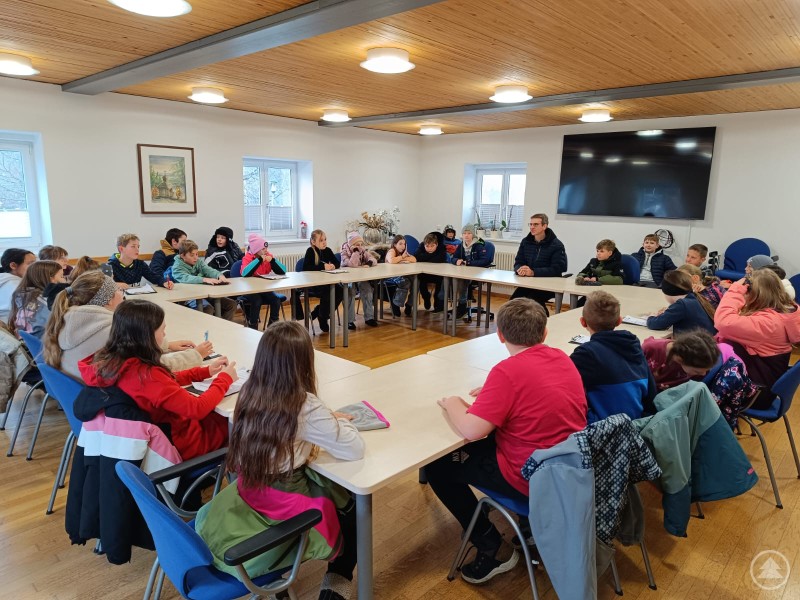 Die Viertklässler befragen im Sitzungssaal Bürgermeister Karlheinz Roth. Eine große Gruppe von Kindern sitzt gemeinsam mit einigen Erwachsenen in einem modernen Sitzungssaal in U-Form um mehrere zusammengestellte Tische. Im Mittelpunkt des Interesses steht eine Person (Bürgermeister) am Kopf der Tischgruppe. Die Kinder hören aufmerksam zu, stellen Fragen oder machen sich Notizen. Im Hintergrund sind Fenster, ein großer Bildschirm und einige Pflanzen zu sehen.