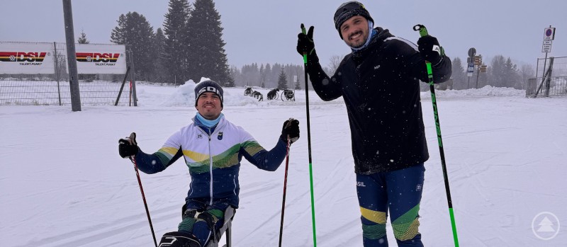 Das Team aus Brasilien hat viel Spaß im Schnee von Finsterau. Zwei Mitglieder eines Teams stehen im Schnee mit Langlaufskiern und lächeln in die Kamera, es schneit leicht.