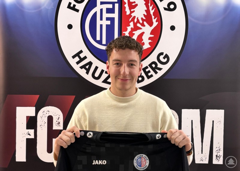 Neuzugang Philipp Reitberger präsentiert sein Trikot beim FC Sturm Hauzenberg. Ein junger Fußballspieler hält ein schwarzes Trikot in die Kamera, im Hintergrund ist das Vereinslogo des FC Sturm Hauzenberg zu sehen.