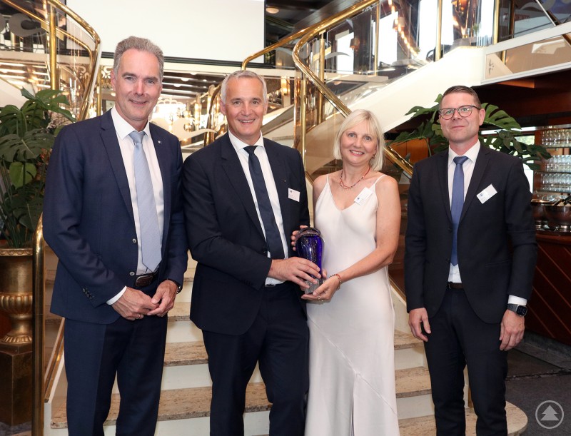 Margit und Florian Noé präsentieren stolz den Cross Border Award. Überreicht wurde ihnen der Preis von IHK-Hauptgeschäftsführer Alexander Schreiner (l.) und IHK-Vizepräsident Jürgen Greipl. Vier Personen in festlicher Kleidung stehen auf einem Schiff vor einer repräsentativen Treppe. Eine Frau in einem weißen Kleid hält gemeinsam mit einem Mann einen blauen Glaspokal. Alle Personen lächeln in die Kamera.