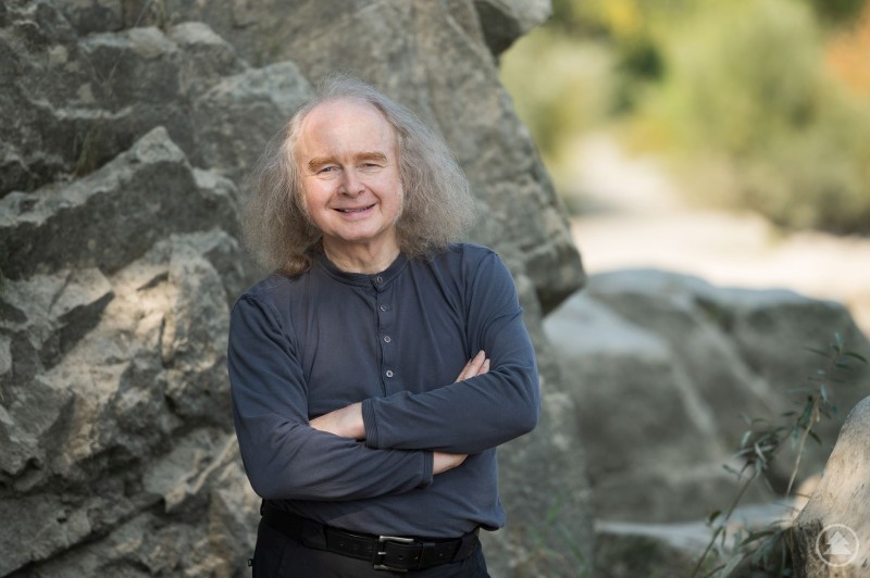 In eine Welt voller Sprachbilder wird Friedrich Hirschl, Kulturpreisträger des Landkreises Passau, am Freitag, den 10. Oktober zusammen mit dem Publikum eintauchen. Ein Mann mit grauem, schulterlangem Haar steht lächelnd mit verschränkten Armen vor einer Felswand in der Natur.
