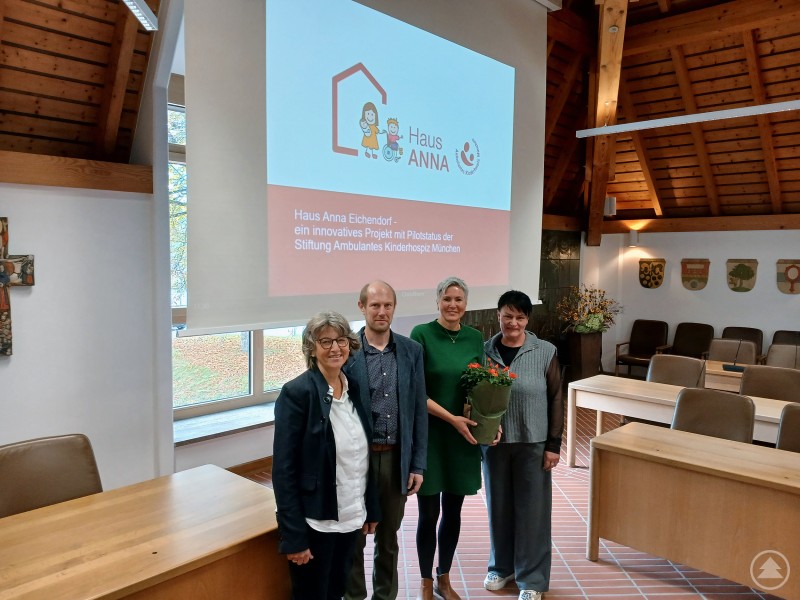 (v. l.) Martina Haintze (KoKi), Thomas Seidl (Amt für Kinder und Familie), Daniela Safajoo (Haus ANNA) und Manuela Schwarz (KoKi) nach der Vorstellung des Kinderhospizes im Landratsamt Freyung-Grafenau. Vier Personen stehen in einem hellen Sitzungssaal vor einer Leinwand, auf der das Logo des Kinderhospizes „Haus ANNA“ Eichendorf zu sehen ist. Eine der Frauen hält einen Blumenstrauß in den Händen.