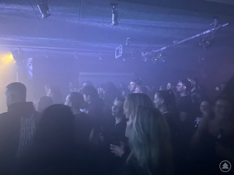 Im dichten Bühnennebel und blauem Licht stehen zahlreiche Menschen dicht gedrängt in einem Club. Die Zuschauer verfolgen gespannt das Konzert, die Atmosphäre wirkt energiegeladen und dicht.