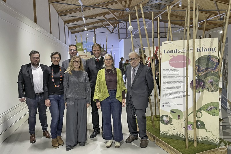 Bezirkstagspräsident Dr. Olaf Heinrich und der wissenschaftliche Leiter der niederbayerischen Freilichtmuseen Dr. Clemens Knobling (hi. Mitte) mit dem Bürgermeister der Gemeinde Mehrere Personen stehen in einer Ausstellungshalle vor einem großen Informationselement mit dem Titel Land.schafft.Klang. Im Hintergrund sind Holzstrukturen und Ausstellungselemente zu sehen.