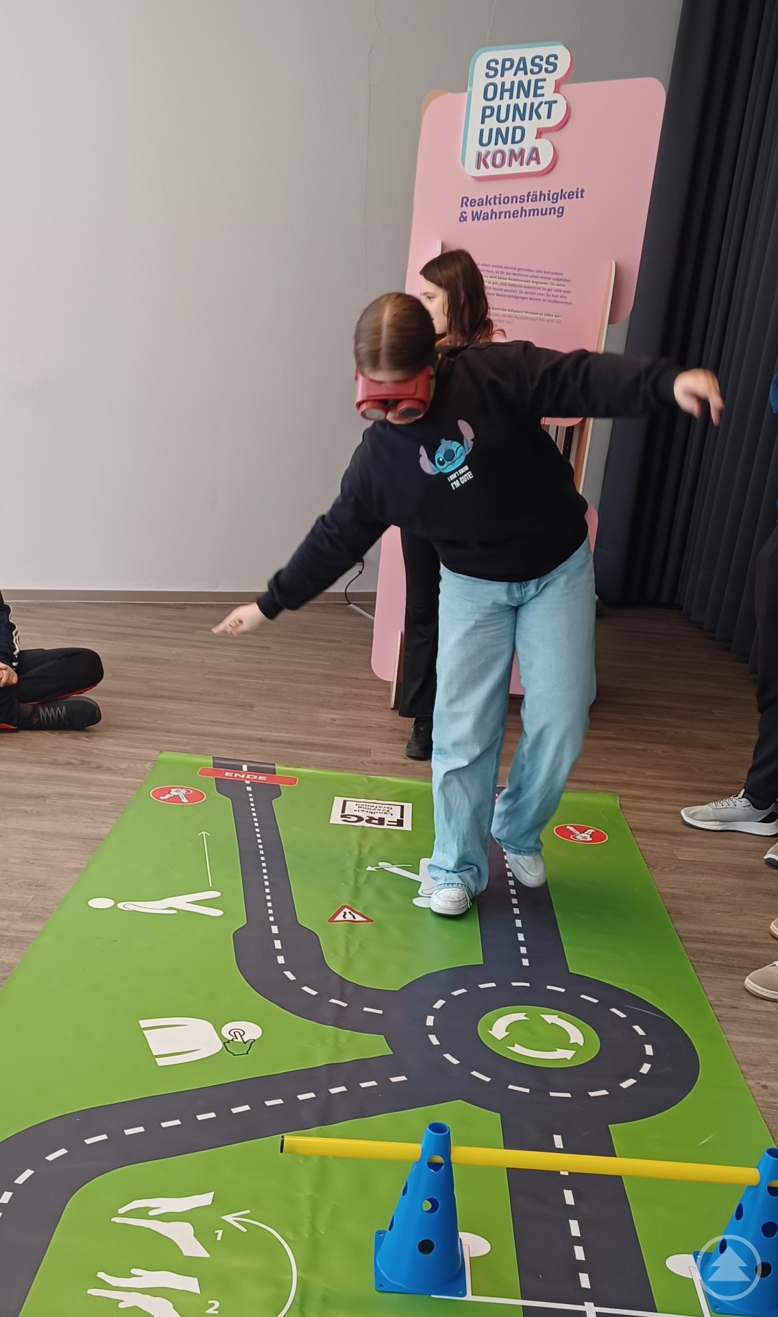 uf dem Bild ist eine Jugendliche zu sehen, die mit Hilfe einer sogenannten „Alkoholbrille“ einen Parcours absolviert. Die Brille simuliert die Wirkung von Alkohol auf die Wahrnehmung und Koordination. Die Teilnehmerin versucht, auf einer geraden Linie zu balancieren, was sichtbar schwerfällt – ein deutliches Zeichen für die Einschränkungen unter Alkoholeinfluss. Im Hintergrund ist ein pinkfarbener Aufsteller mit dem Logo der Ausstellung „Spaß ohne Punkt und Koma“ zu sehen, der die Themen Reaktionsfähigkeit und Wahrnehmung behandelt. Weitere Jugendliche beobachten die Übung.