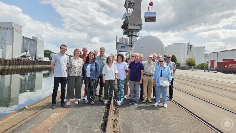 Die Teilnehmer des Ausflugs am Umschlagplatz des Hafens Straubing-Sand, dritter von links Günther Achatz. Gruppe von rund 20 Personen steht auf einem Hafengelände direkt an einem Hafenbecken mit Wasser. Im Hintergrund sind Silos, Tanks, Industriegebäude und ein großer Umschlagkran zu sehen. Es ist leicht bewölkt, aber freundlich.